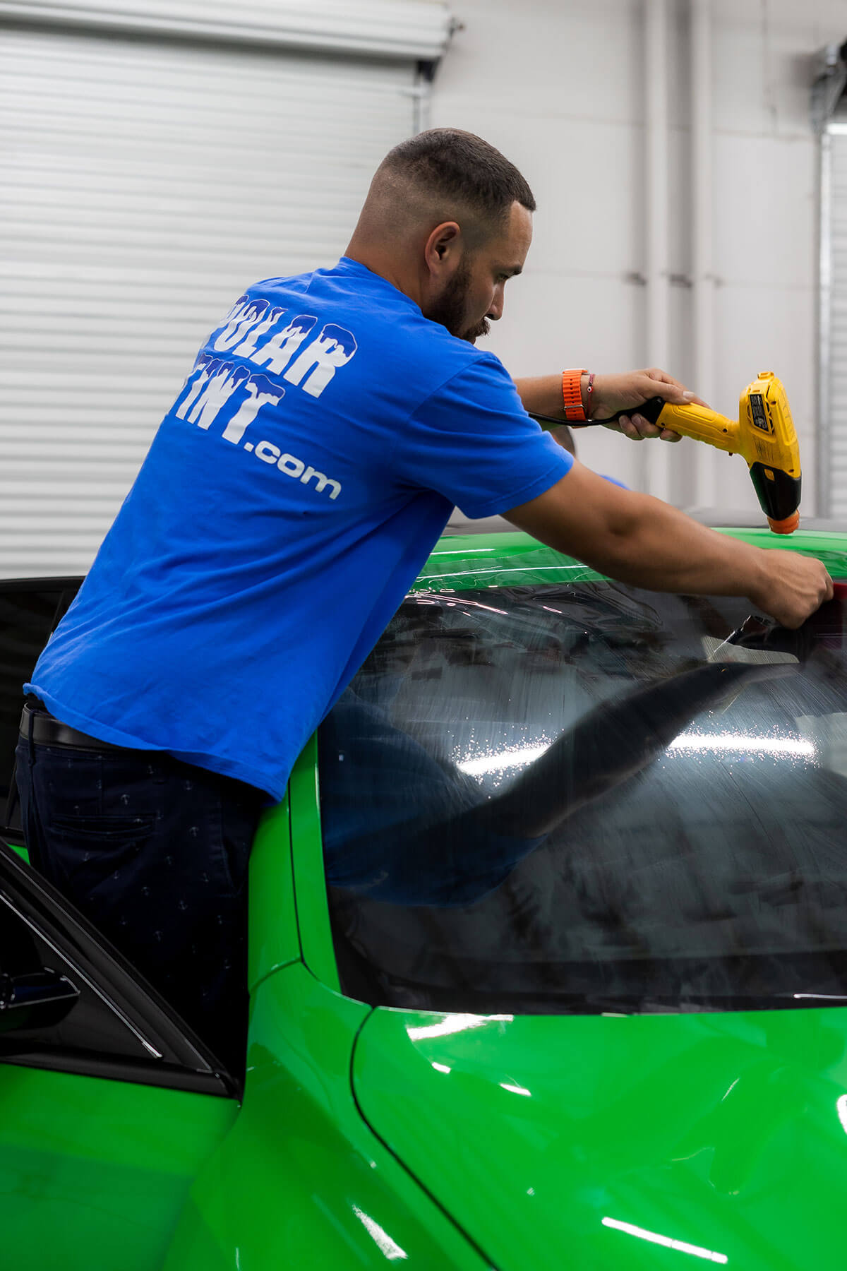 Polar Tint certified installer heat-shrinking nano ceramic window tint film on a vehicle windshield — branded shop tee, in-bay install