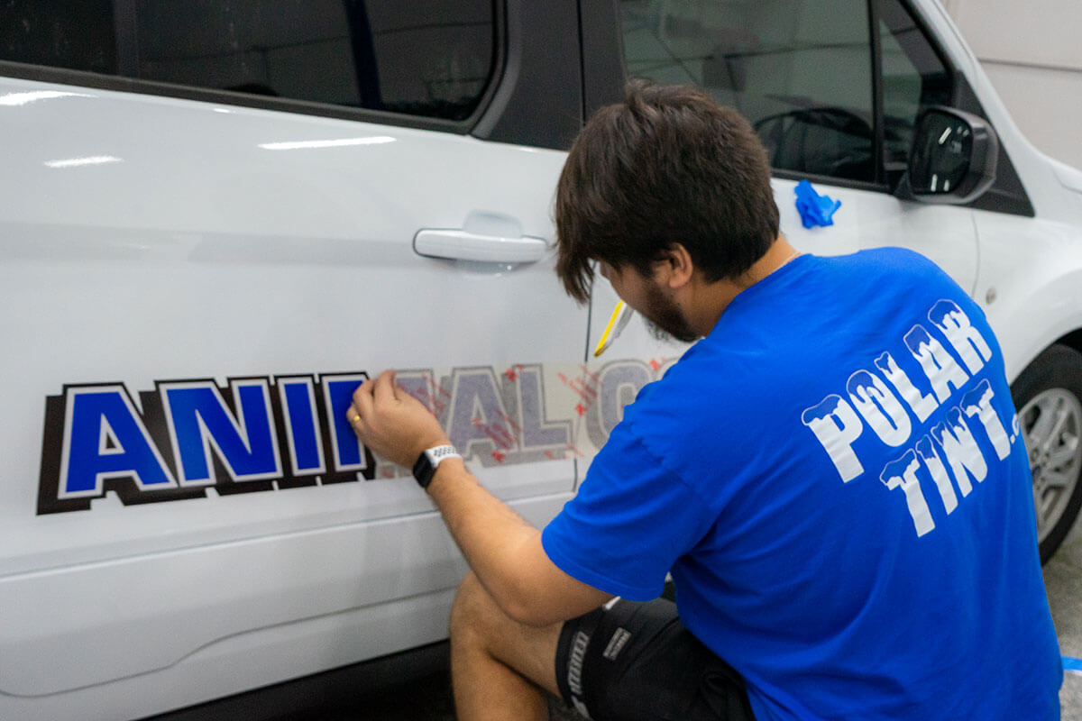 Polar Tint Certified technician in branded shop tee applying custom cut-vinyl door lettering to a white vehicle — in-bay install of door-logo decals